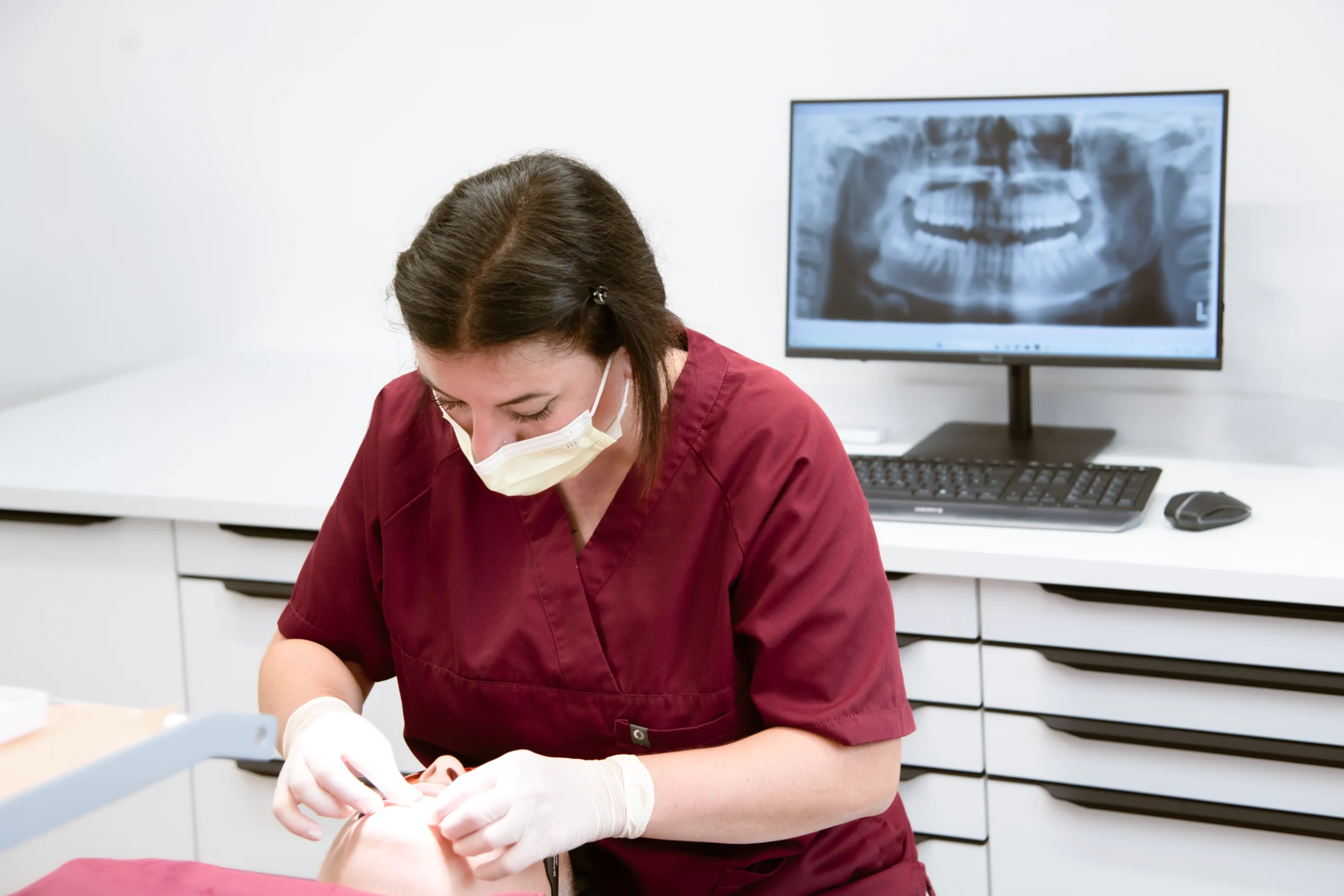 Behandlung einer Patientin durch eine Mitarbeiterin in der kieferorthopädischen Praxis mit digitalem Röntgenbild im Hintergrund.
